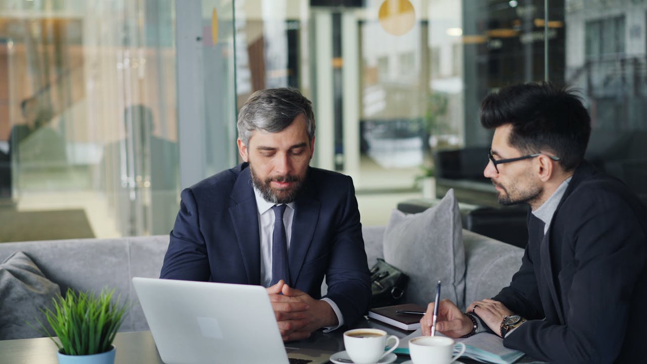 who-we-are Two businessmen in suits discussing work at a cafe with laptops and coffee.