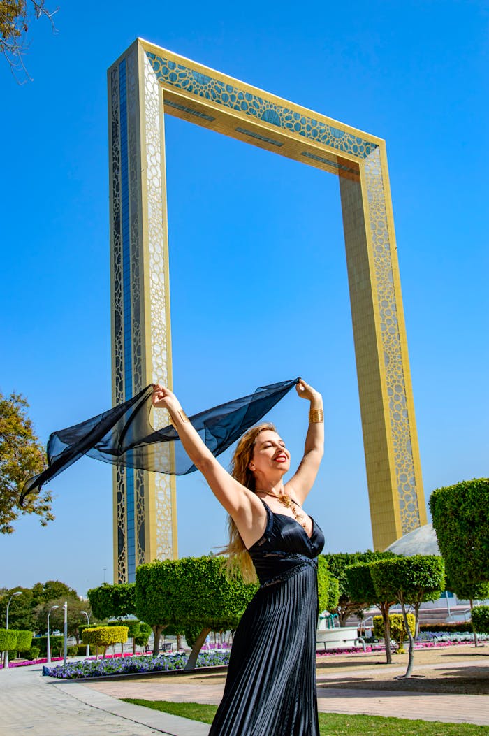 A cheerful woman poses with a scarf under the iconic Dubai Frame on a bright sunny day.
